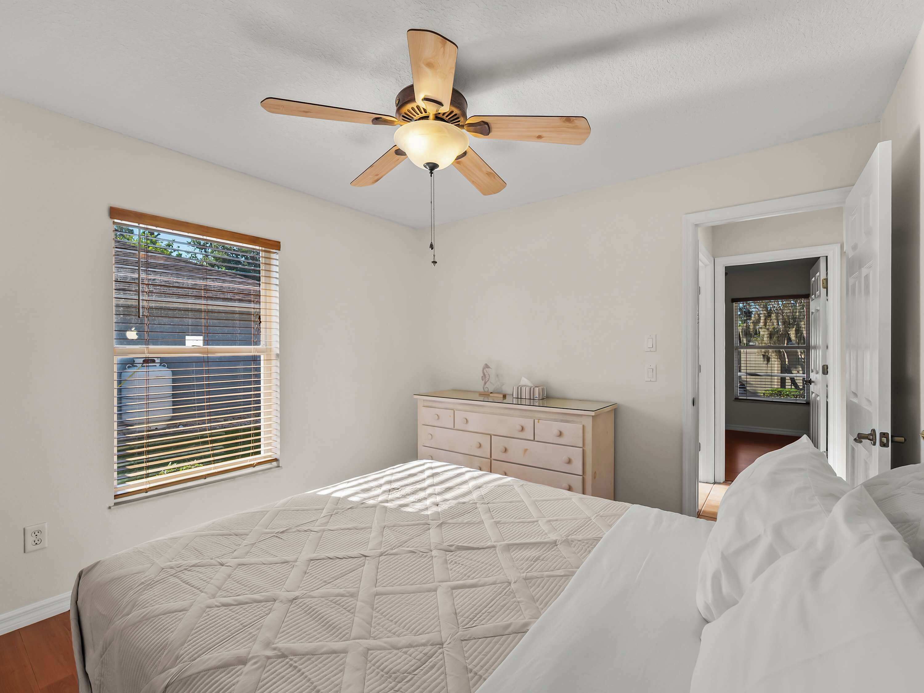 Ein Schlafzimmer mit Bett, Kommode, Deckenventilator und einem Fenster mit Jalousien.