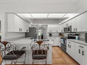 Eine moderne Küche mit weißen Schränken, einer zentralen Kochinsel, zwei verzierten Stühlen und Blick durch einen Bogen.