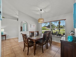 A dining area with a glass table and wooden chairs, a hanging light fixture, and a view through a window with blinds. - Sunlit & Spacious Solar-Heated Screened Pool, BBQ (North Port)