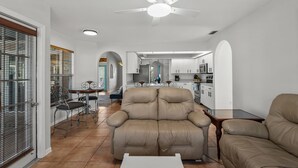 En moderne stue med en beige sofa, et spiseområde med bord og stole, og et køkken i baggrunden.