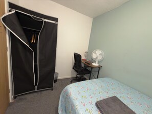 Un dormitorio con cama, un escritorio con ventilador, una silla y un armario.