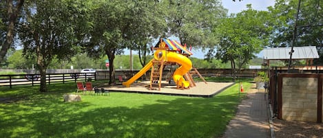 A playground with a slide and climbing structure, surrounded by trees and a fenced area.