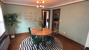 A dining area with a round wooden table, green chairs, a black and white striped rug, a potted plant, and a ceiling light fixture.