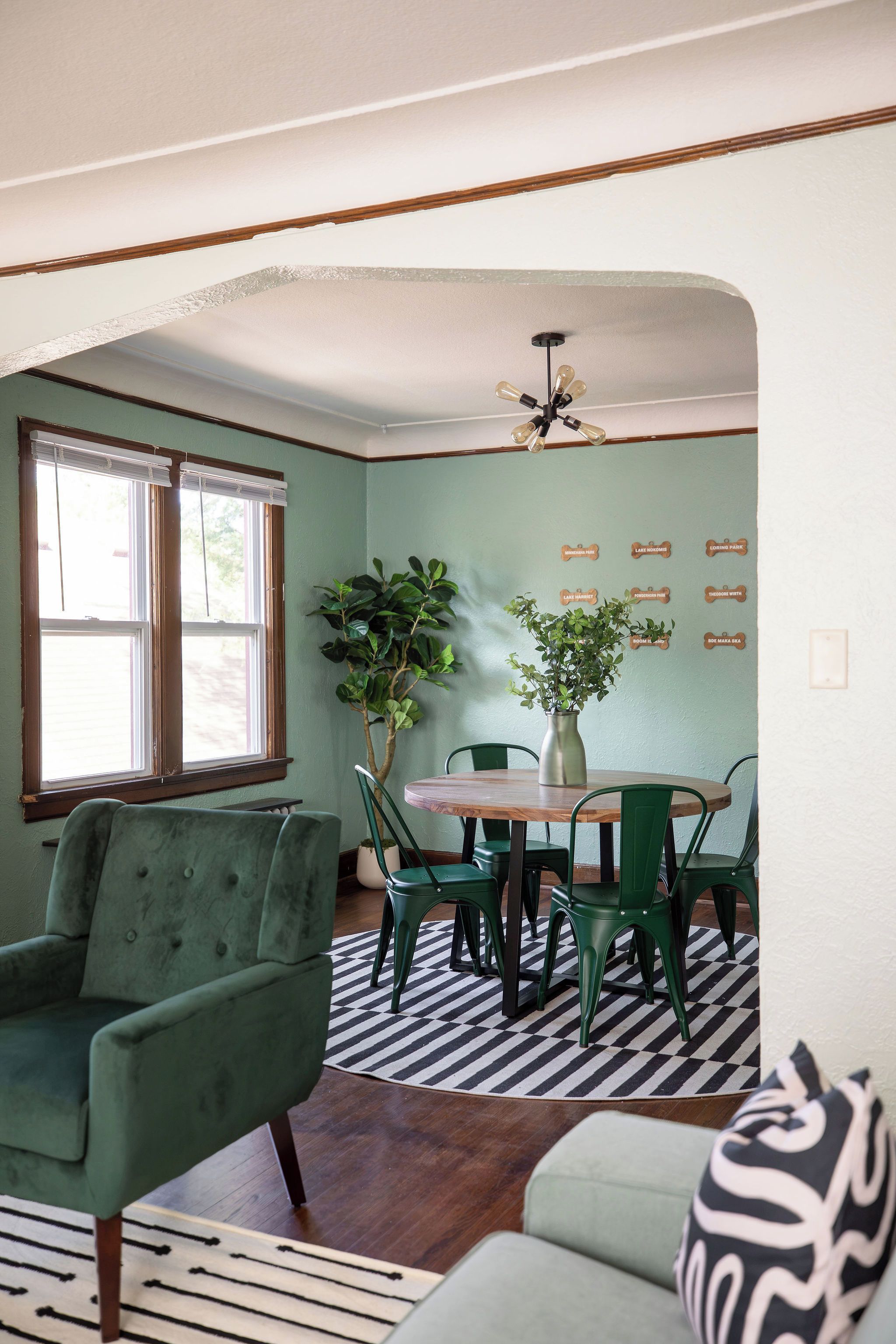 Un comedor con mesa de madera, sillas verdes, una planta en maceta y un tapete de cuadros blancos y negros.