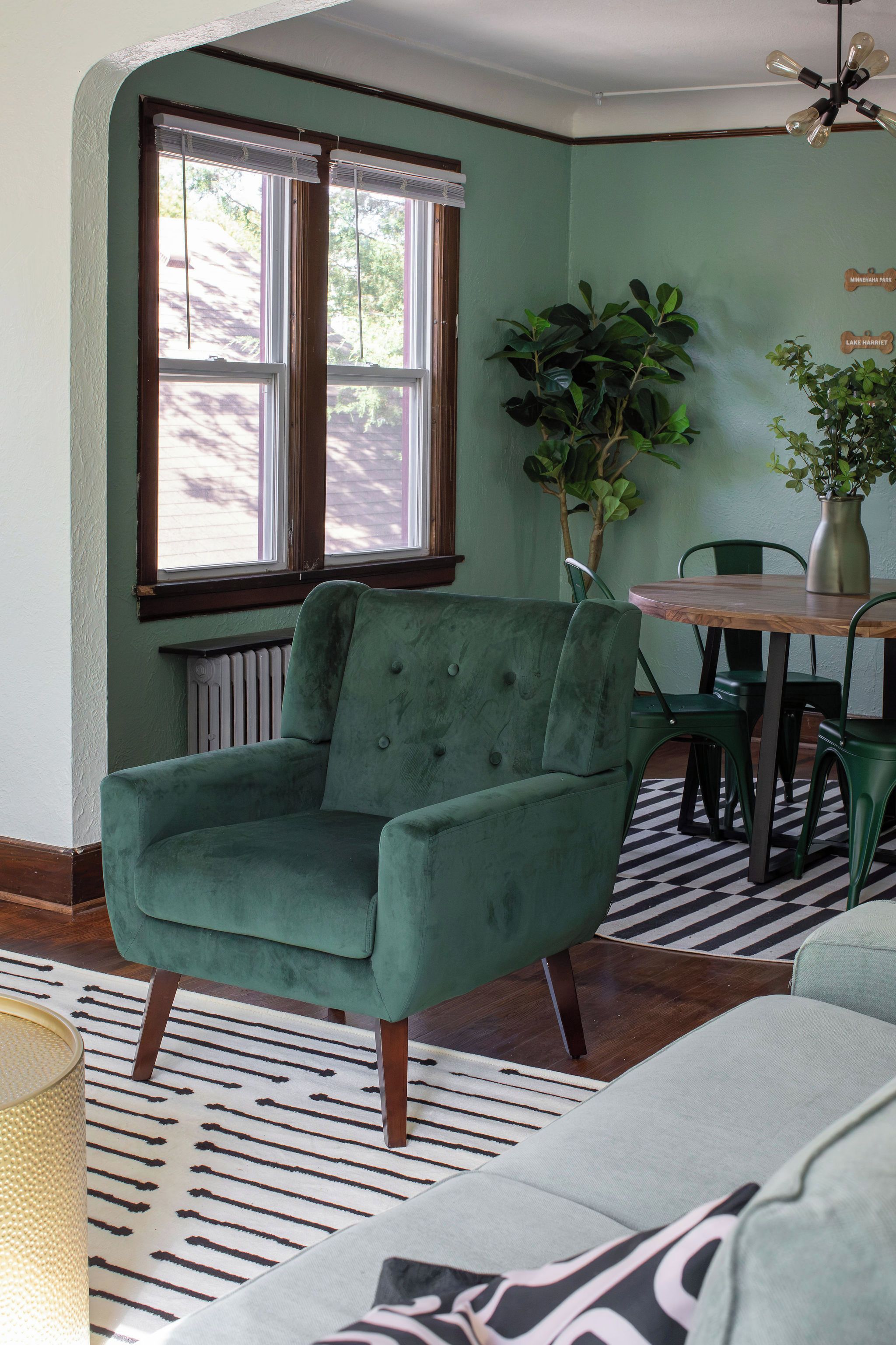 Una habitación con un sillón verde, una mesa de comedor con sillas, una planta en maceta y una ventana con persianas.