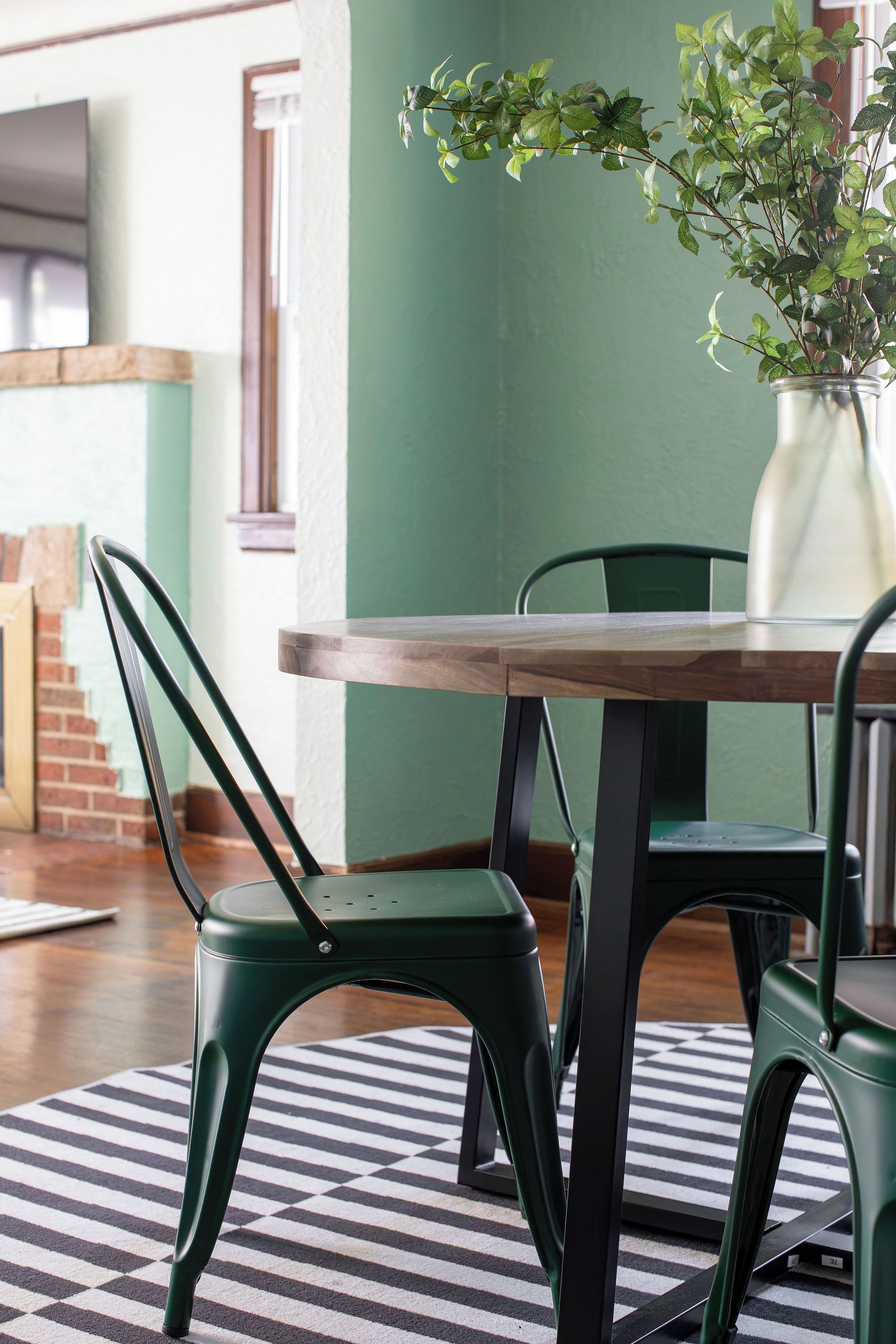 A dining area with a wooden table and green chairs, a striped rug, and a potted plant.