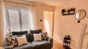A modern living room with a grey sofa, patterned cushions, a wooden floor, a chandelier, and a small side table with plants. - Stylish 2-Bed Bedford Home | Sleeps 4 + Sofa Bed | Free Parking |Super Fast WiFi (Bedford)