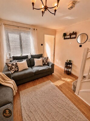 A modern living room with a grey sofa, patterned cushions, a wooden floor, a chandelier, and a small side table with plants. - Stylish 2-Bed Bedford Home | Sleeps 4 + Sofa Bed | Free Parking |Super Fast WiFi (Bedford)