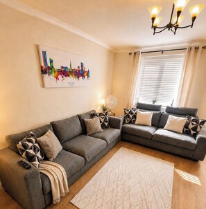 Ein Wohnzimmer mit zwei grauen Sofas, einem beigen Teppich, einem Stadtbildgemälde, einer Deckenleuchte und einem Fenster mit Vorhängen.