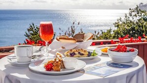 A table set with a glass of red beverage, a cup, a plate with berries, a bowl of fruit, and a menu on a sunny day with a view of the sea.