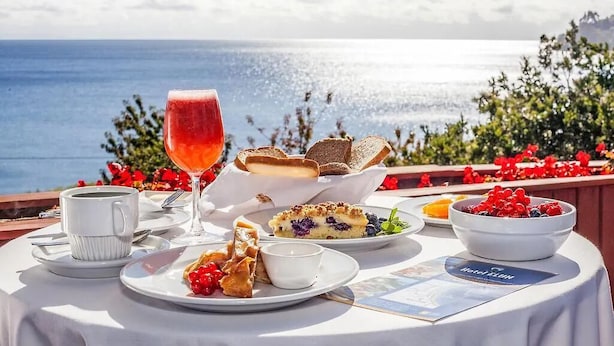 Una mesa dispuesta con un vaso de bebida roja, una taza, un plato con bayas, un bol de fruta y un menú, todo bajo un sol radiante con vistas al mar.