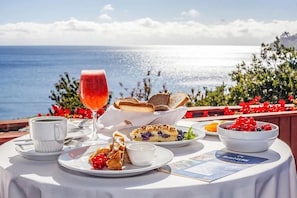 A table set with a glass of red beverage, a cup, a plate with berries, a bowl of fruit, and a menu on a sunny day with a view of the sea. - Casa Freude Hotel Boutique (Frutillar)