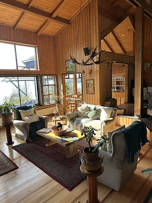 A spacious living room with wooden walls and a high ceiling, featuring a large window, a fireplace, and a bar area.