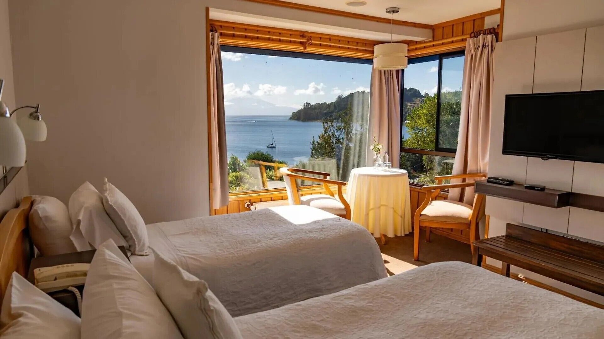 A hotel room with two beds, a TV, a table with chairs, and a view of the sea.