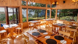 Interior de madera con varias mesas y sillas de comedor, amplias ventanas con vistas a zonas verdes.