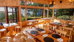Interior de madera con varias mesas y sillas de comedor, amplias ventanas con vistas a zonas verdes.