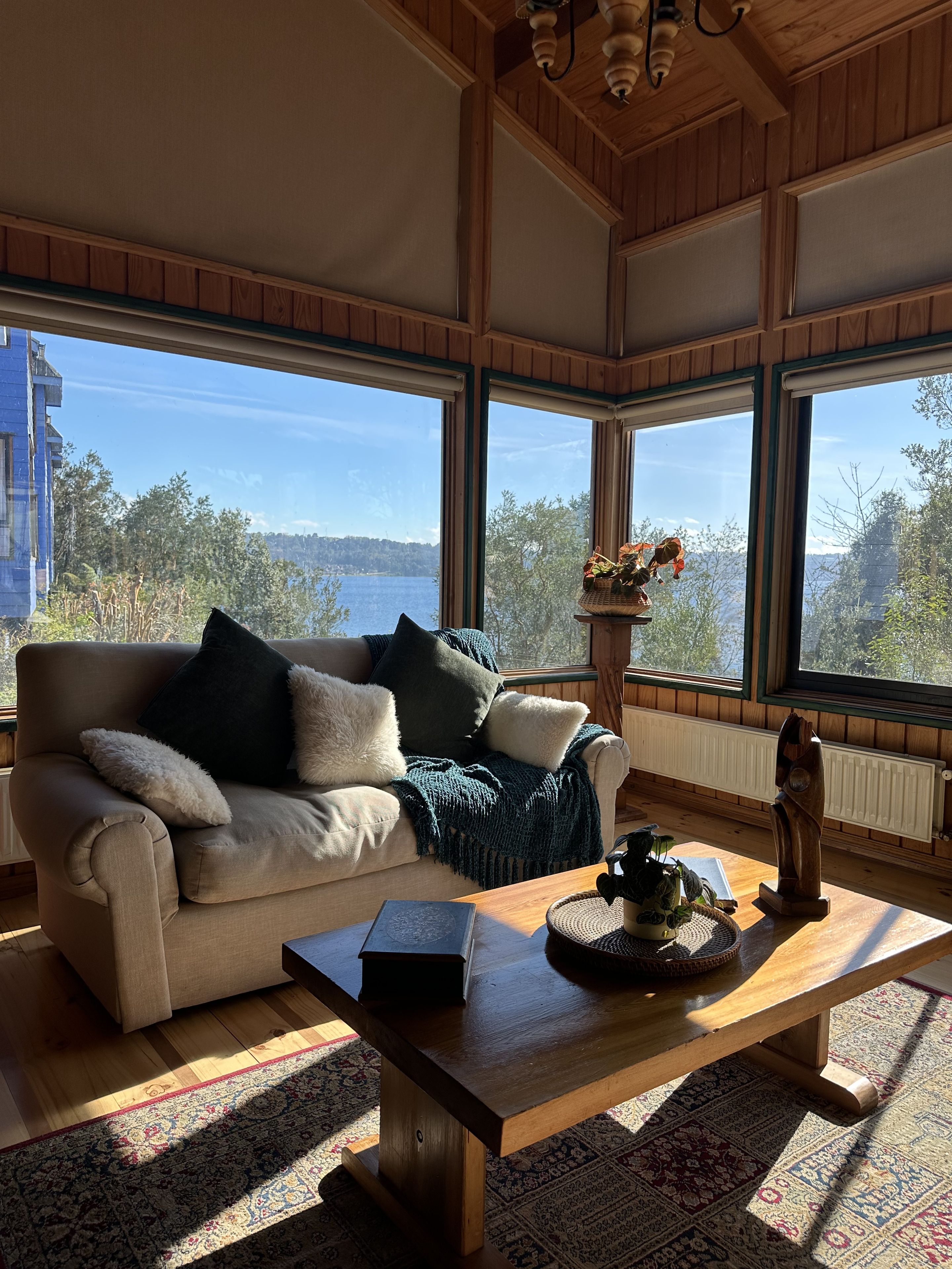A cozy room with a wooden ceiling, a beige sofa with cushions, a wooden coffee table, and a view of a lake through large windows.