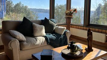 A cozy room with a wooden ceiling, a beige sofa with cushions, a wooden coffee table, and a view of a lake through large windows.