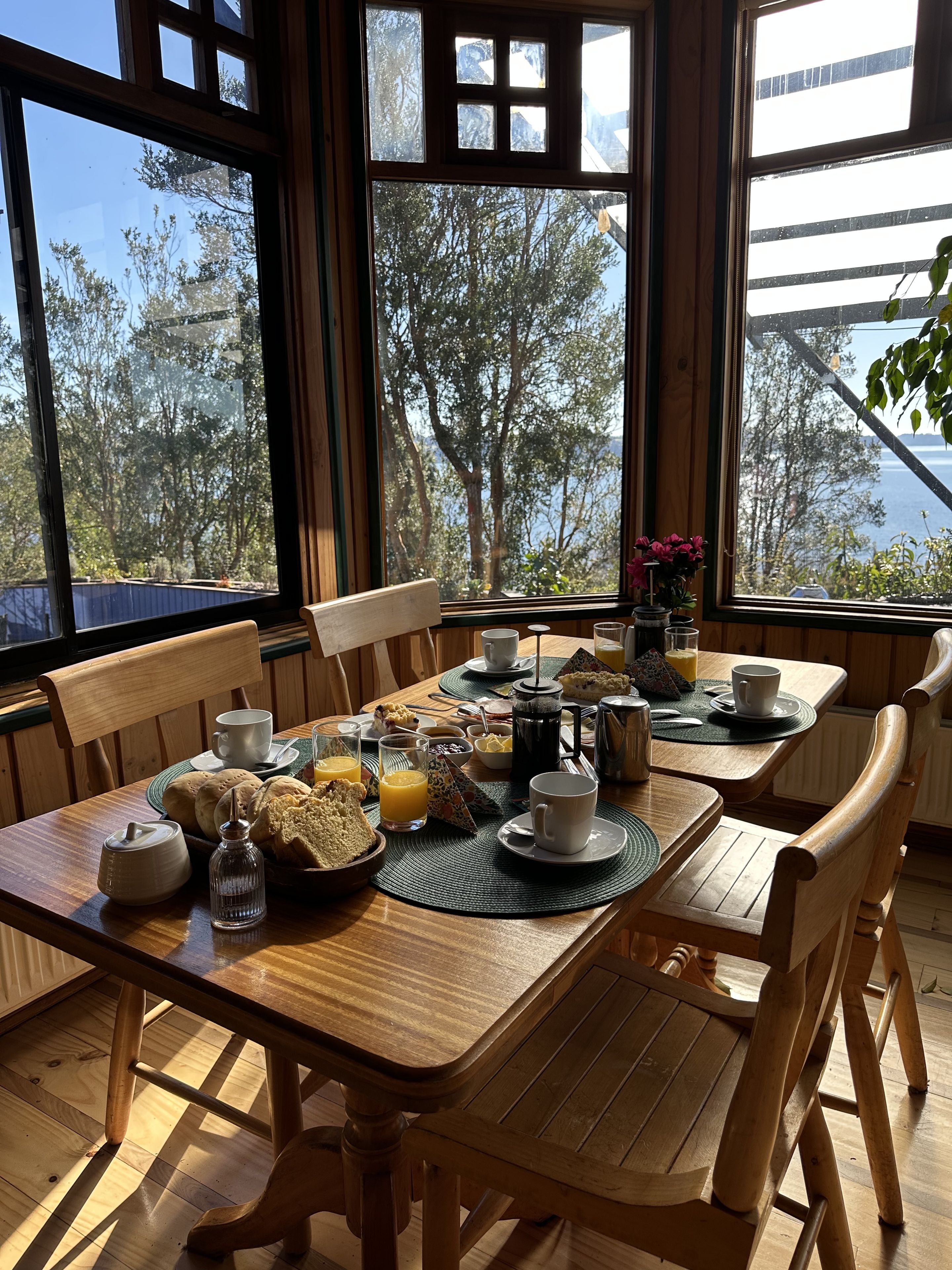 A wooden dining table with breakfast items, two chairs, and large windows with a view of trees and a body of water.