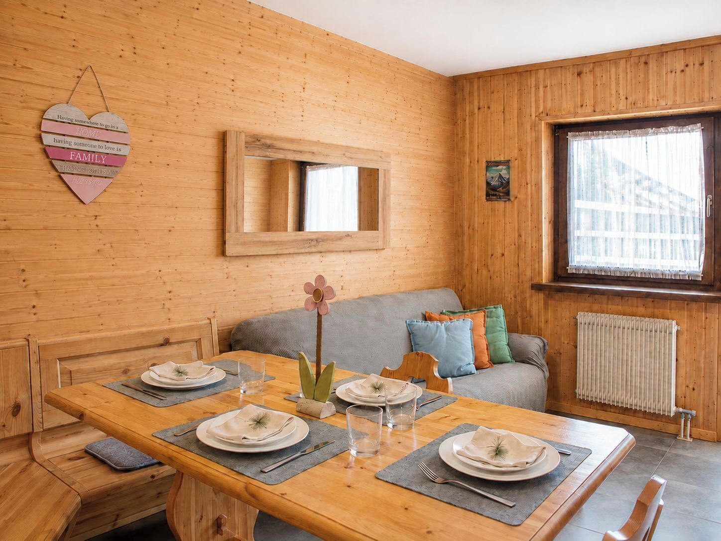 Ein Holzinnenraum mit einem für vier Personen gedeckten Esstisch, einer Couch mit Kissen und einer herzförmigen Dekoration an der Wand.