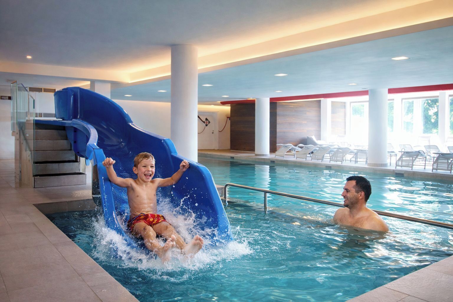 Ein Hallenbad mit blauer Rutsche, einer Treppe und Liegestühlen.