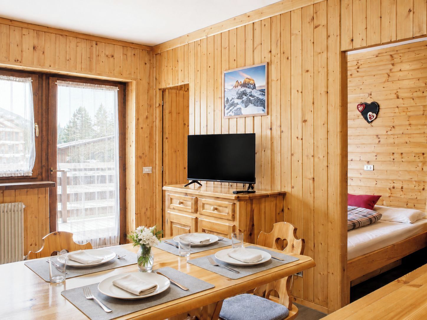 Ein Zimmer mit Holzwänden, einem für zwei Personen gedeckten Esstisch, einem Fernsehgerät auf einem Schrank und einem Bett mit einem roten Kopfkissen.