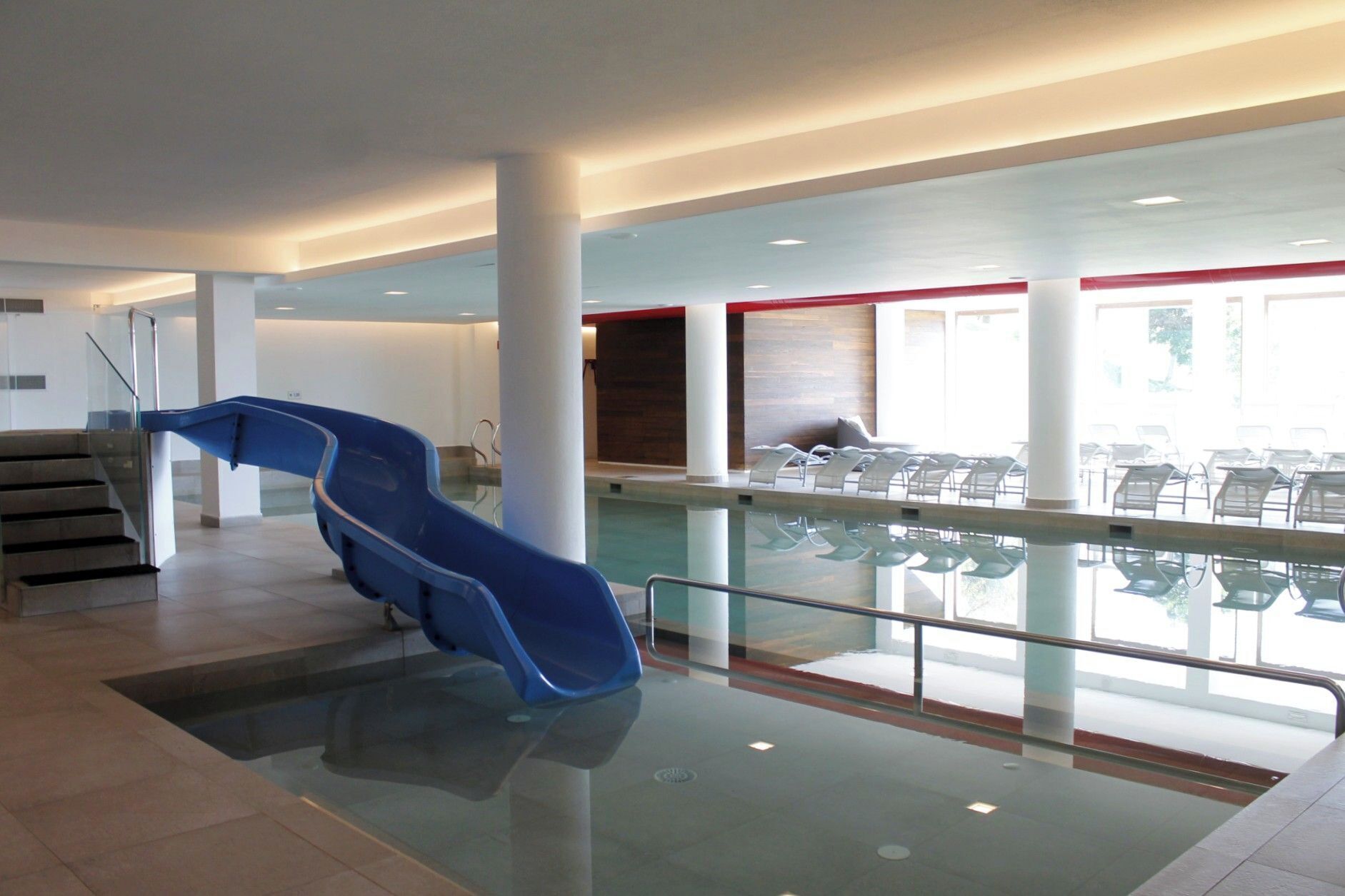 Zona d'una piscina interior moderna, amb un tobogan blau, una escala i seients al costat de la piscina.