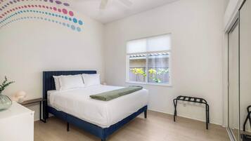 Quarto com cama, janela com vista para o verde, ventilador de teto e mesa de cabeceira com abajur.