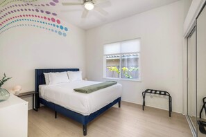 Un dormitorio con cama, ventana con vista a follaje, ventilador de techo y mesita de noche con lámpara.