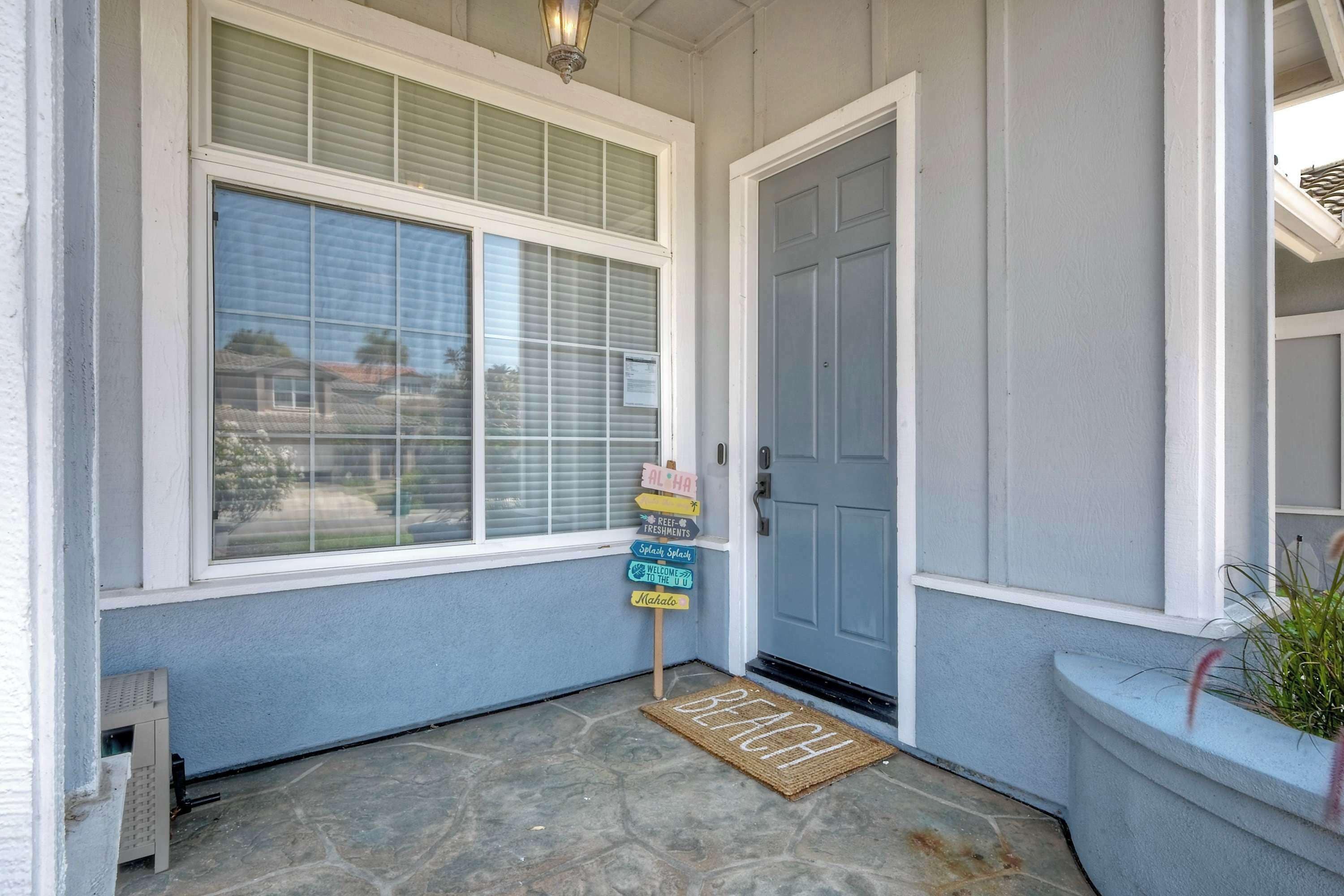 Uma entrada de casa com porta azul, janela com persianas, tapete de boas-vindas e placa com diversas mensagens.