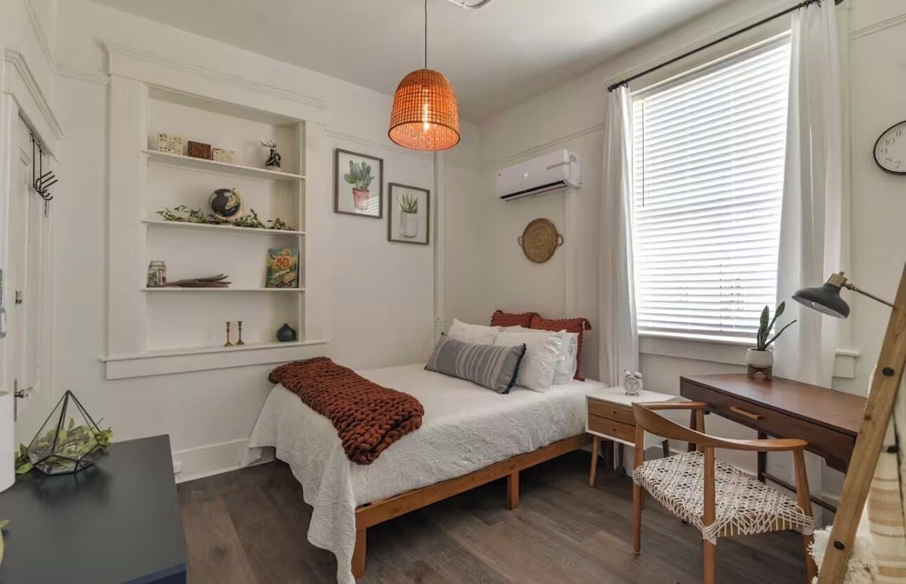 Un dormitorio con cama, mesita de noche, silla, estantes y una ventana con persianas.