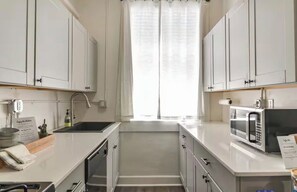 A modern kitchen with white cabinetry, a stainless steel sink, a microwave, and a window with sheer curtains. - Apartment in Historic Downtown Prescott - Fremont Room (Prescott)