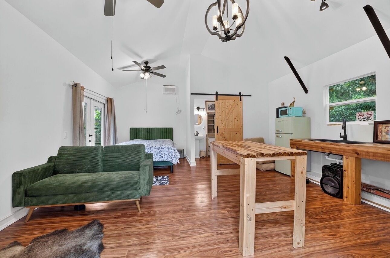 Una habitación con sofá verde, una mesa de madera, un ventilador de techo y una ventana con vista a follaje.