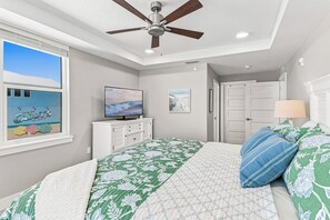 Un dormitorio con cama, ventilador de techo, televisor, ventana que muestra una playa y una puerta.