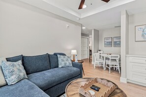 A modern living room with a sofa, a dining area with a table and chairs, and a kitchen area with cabinets. - Beach Flower 201 - Beautiful Beachfront Condo w/ Balcony, Elevator, & Beach Gear. (Madeira Beach)
