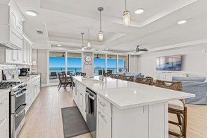 A modern kitchen with a large island, dining area with wicker chairs, and a view of the ocean. - Beach Flower 201 - Beautiful Beachfront Condo w/ Balcony, Elevator, & Beach Gear. (Madeira Beach)