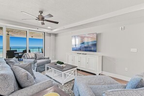 A modern living room with a view of the ocean, a ceiling fan, and a flat-screen TV on a white cabinet. - Beach Flower 201 - Beautiful Beachfront Condo w/ Balcony, Elevator, & Beach Gear. (Madeira Beach)