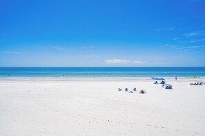 A beach with white sand, clear blue water, and a few umbrellas and chairs. - Beach Flower 201 - Beautiful Beachfront Condo w/ Balcony, Elevator, & Beach Gear. (Madeira Beach)