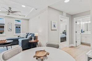 A modern living room with a round table, a sofa, a ceiling fan, and a view of the sea through the windows. - Beach Flower 201 - Beautiful Beachfront Condo w/ Balcony, Elevator, & Beach Gear. (Madeira Beach)