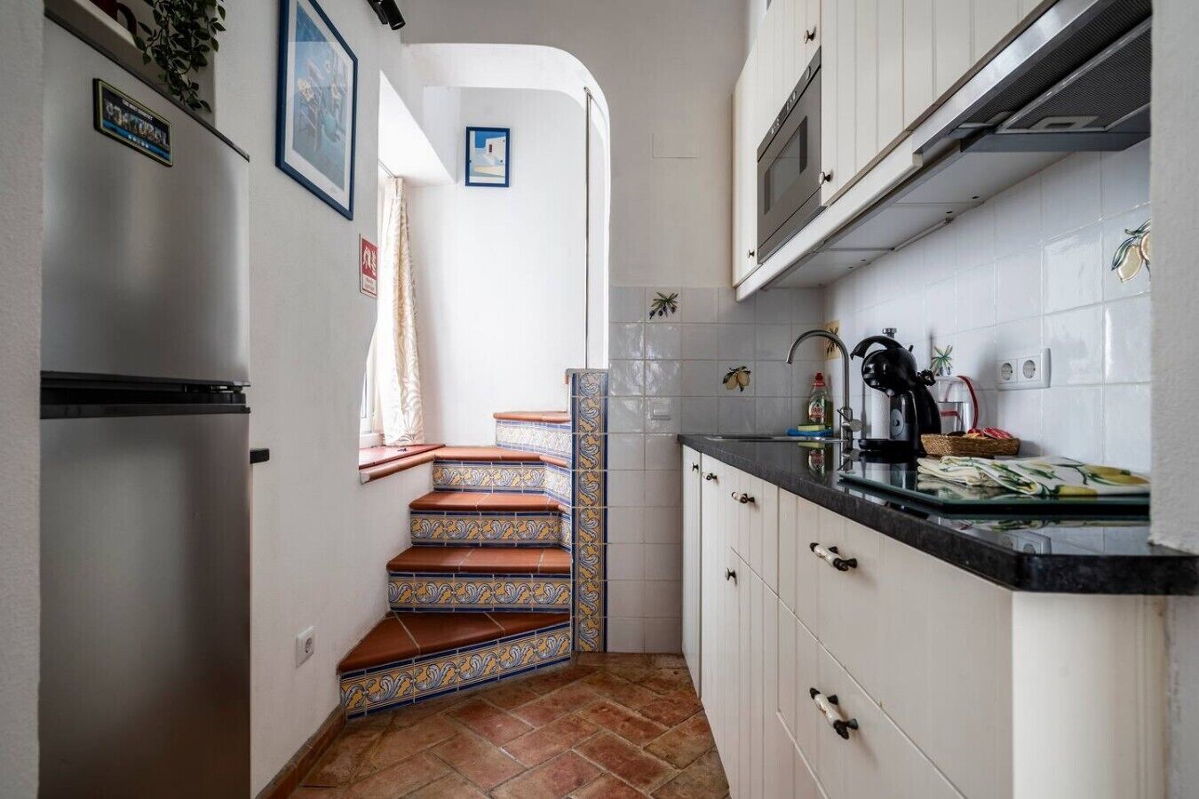 Una cucina con mobili bianchi, un frigorifero in acciaio inossidabile, una parete rivestita di piastrelle e un piano di lavoro nero.