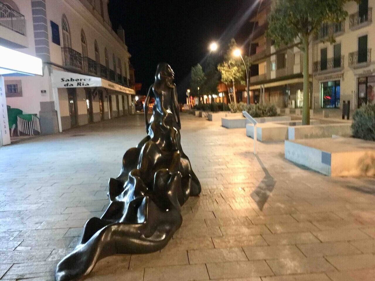Une sculpture se trouve sur une place publique, avec des bâtiments en arrière-plan.