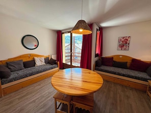 Una habitación con dos sofás de madera, una mesa redonda de madera, una lámpara colgante, un espejo y una ventana con cortinas rojas.