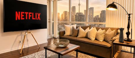 Una sala de estar con un televisor grande mostrando 'NETFLIX', un sofá con cojines, una mesa de centro, una lámpara y vistas a un horizonte urbano.
