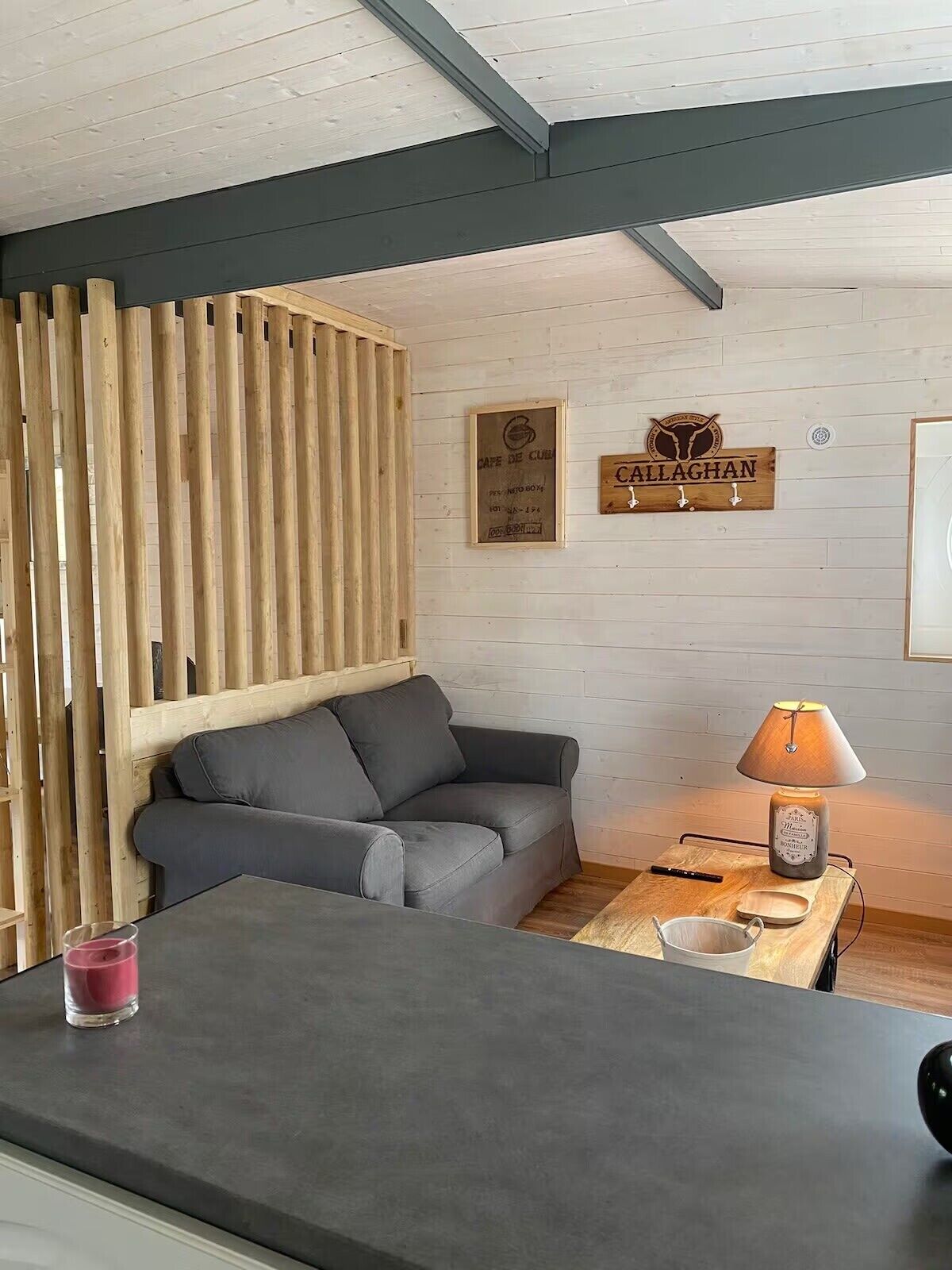 A room with a grey sofa, a wooden partition, a table with a lamp, and two signs on the wall.