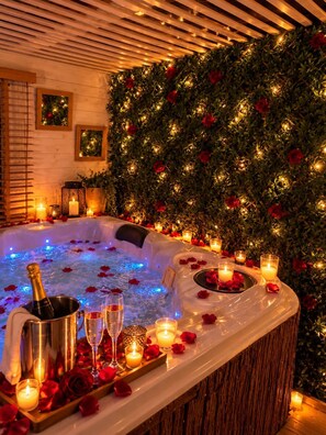 A hot tub surrounded by candles and rose petals, with a wooden wall and greenery in the background.