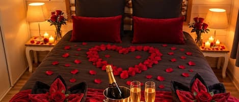 Un lit décoré de pétales de roses disposés en forme de cœur, deux oreillers rouges et un plateau avec une bouteille de champagne, des verres et des bougies.