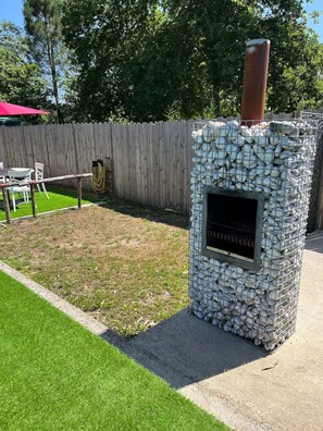 Una parrilla de piedra al aire libre con chimenea, rodeada por una valla de madera y un césped.