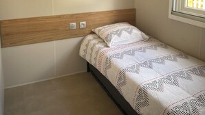 A single bed with a patterned bedspread, a wooden headboard, a wall-mounted shelf, and a window with daylight. - Mobil-home 3 chambres 1 sdb (Mimizan)