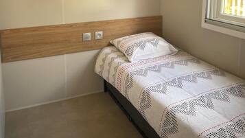 A single bed with a patterned bedspread, a wooden headboard, a wall-mounted shelf, and a window with daylight.