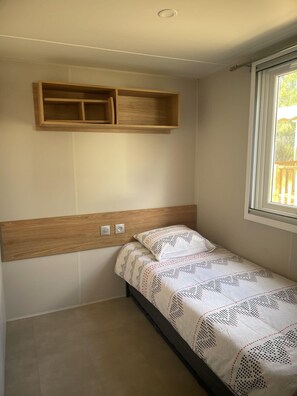 Ein Einzelbett mit gemustertem Überwurf, ein Holzkopfende, ein Wandregal und ein Fenster mit Tageslicht.